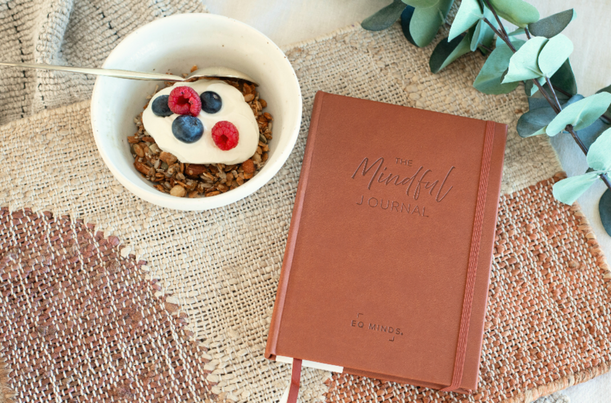 Brown journal by EQ Minds titled 'The Mindful Journal' on a textured surface with a bowl of yogurt and berries.